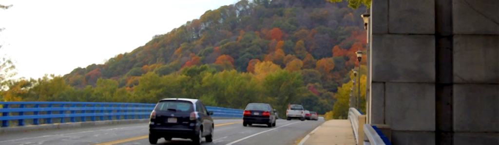 A bridge with cars heading toward a mountain covered in trees with fall foliage.